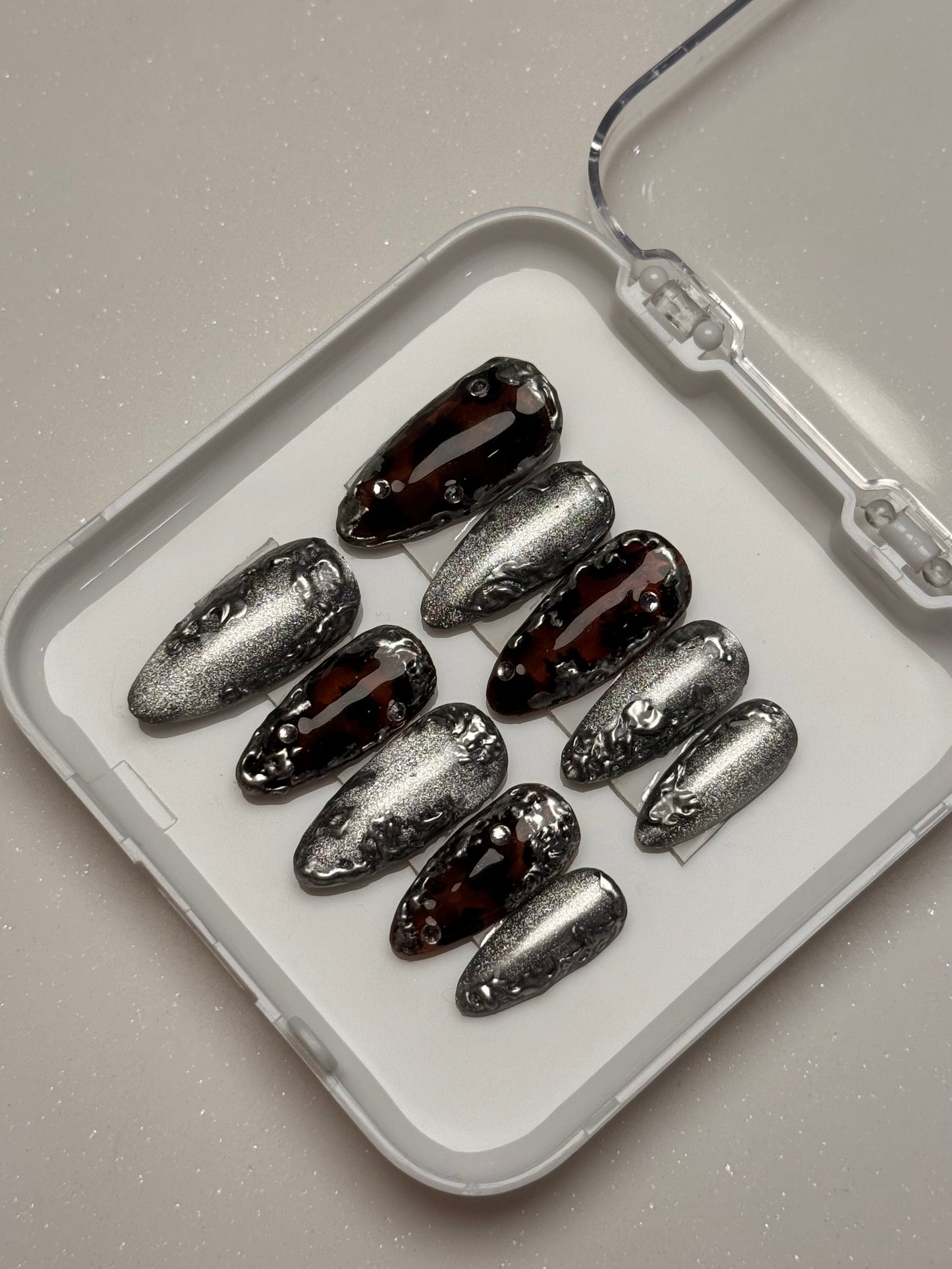 Set of silver cat eye and dark brown nail art tortoise shell nail set components on a white tray with a clear lid.