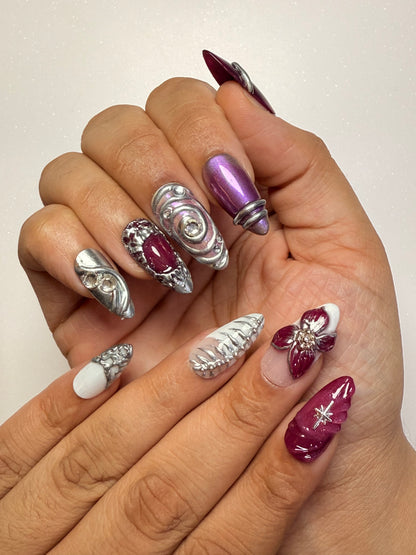 Hand with decorative nails featuring purple, silver, and white designs on a plain background chrome nail set 3D flower nail set  purple nail set