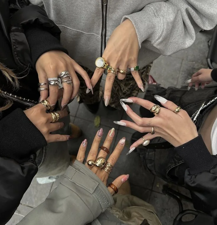 Four hands stacked in a circle, each with long press-on nails and gold and silver rings