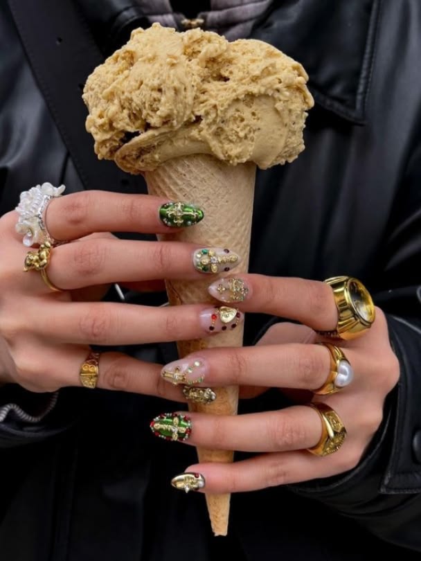 Hand with green and gold botanical 3D nail art holding a caramel ice cream cone, gold rings stacked on every finger